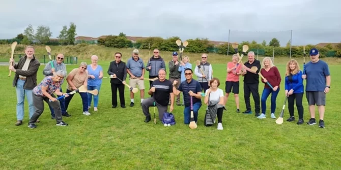 a group of adults posing on a grass field while participating in a hurling experience, a popular cultural activity in Ireland.